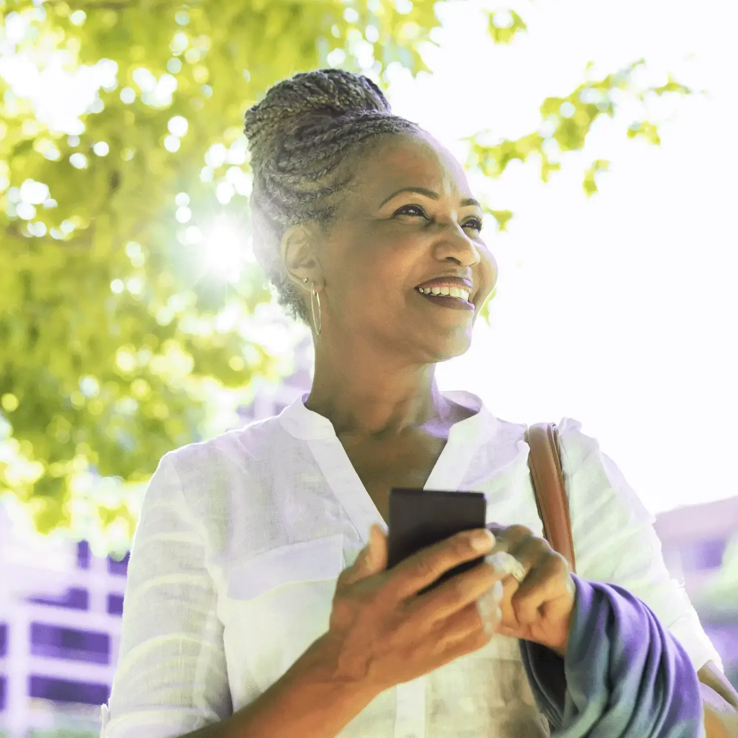 Mulher 50+ sorrindo ao ar livre com um celular na m&atilde;o, sob a ilumina&ccedil;&atilde;o do sol entre as &aacute;rvores.