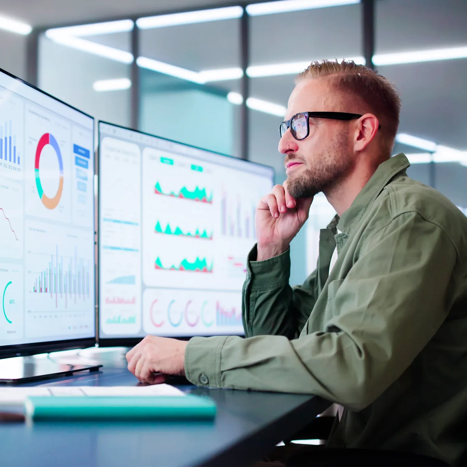 Profissional de &oacute;culos analisando gr&aacute;ficos em dois monitores de computador.